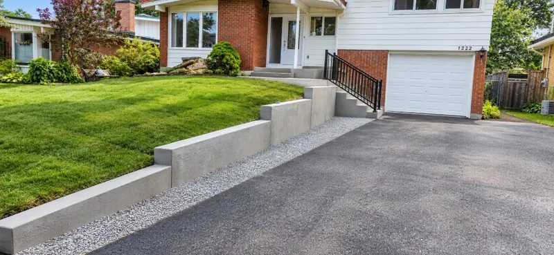 Front yard with a stepped concrete retaining wall, concrete steps and a black railing beside the driveway